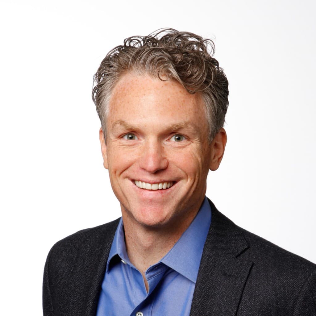 Professional headshot of Nathan smiling, salt-and-pepper hair, light blue shirt and dark blazer, on a clean white background.